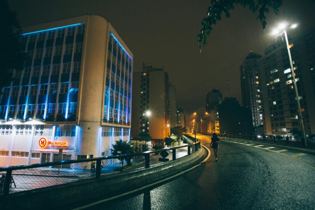A solitary person walks on a wet city street at night, surrounded by illuminated buildings.
